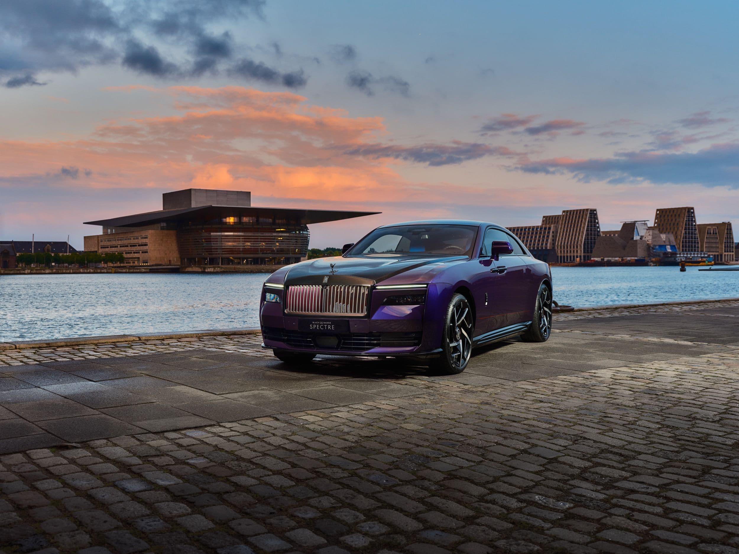 A luxurious purple Rolls-Royce coupe parked by the water with modern architecture in the background.
